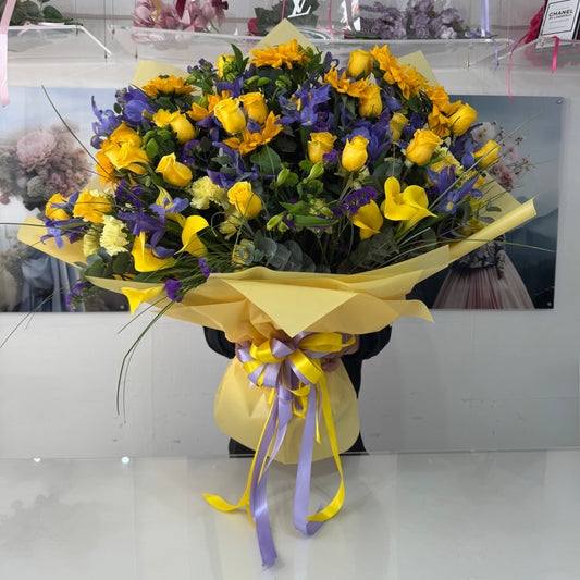 Bouquet of yellow and purple flowers with a yellow wrap and purple ribbon on a white surface.